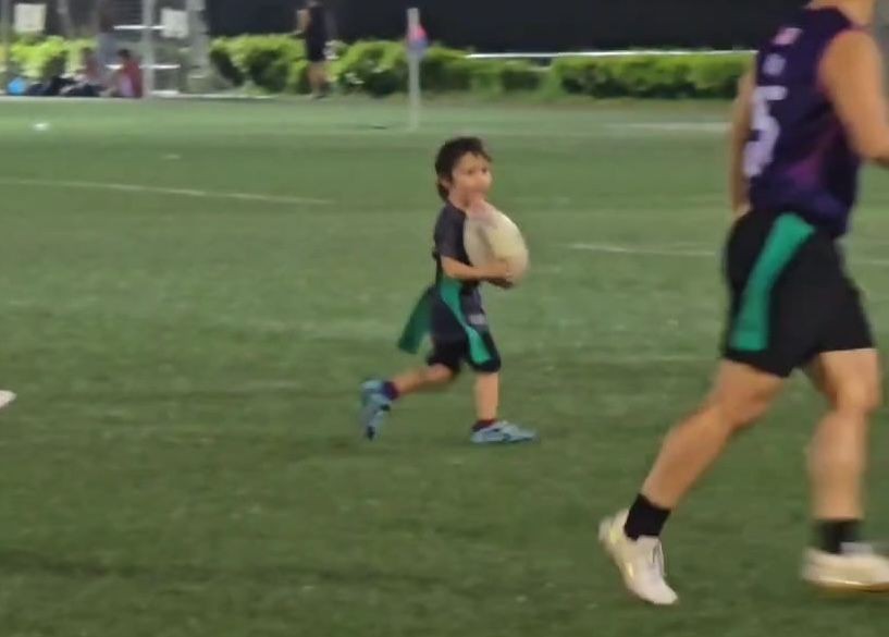 Young child holding a rugby ball on a field, with an adult player nearby.