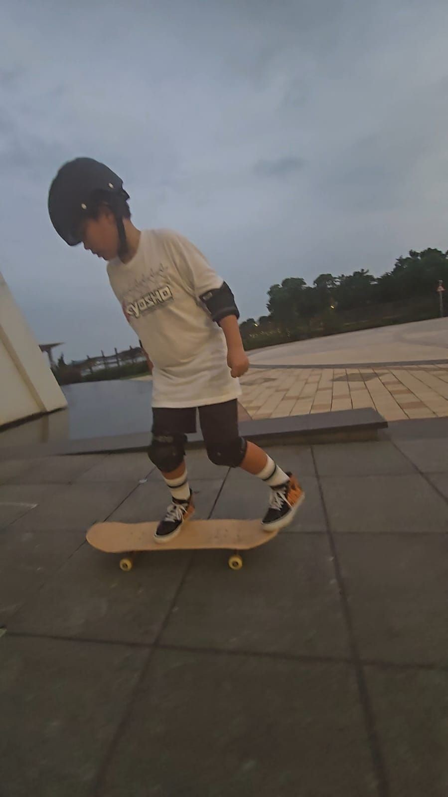 Young child wearing safety gear skating on a skateboard in an outdoor plaza during a cloudy day.