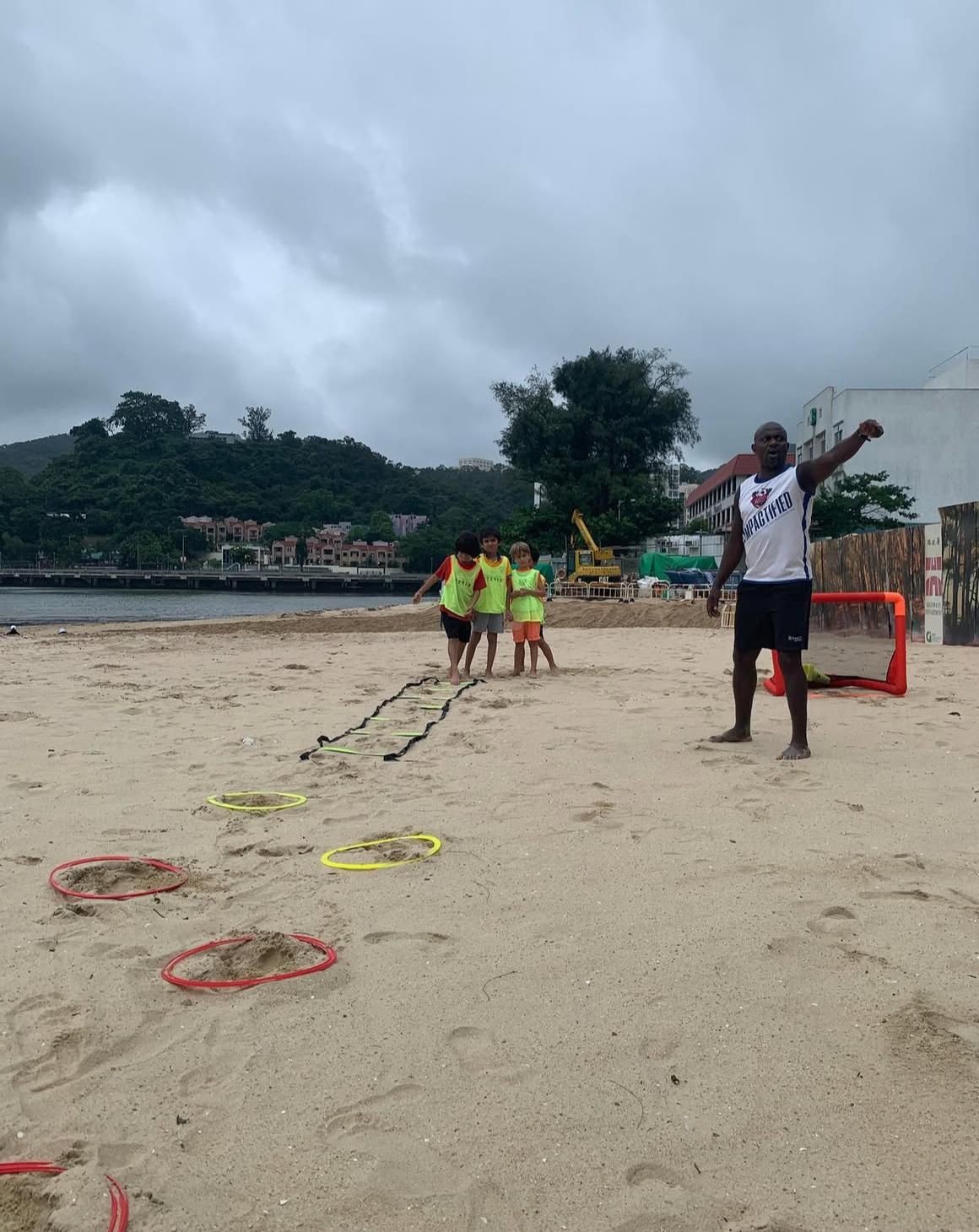 Children and coach playing a game on a sandy beach with agility rings and ladder under cloudy skies.