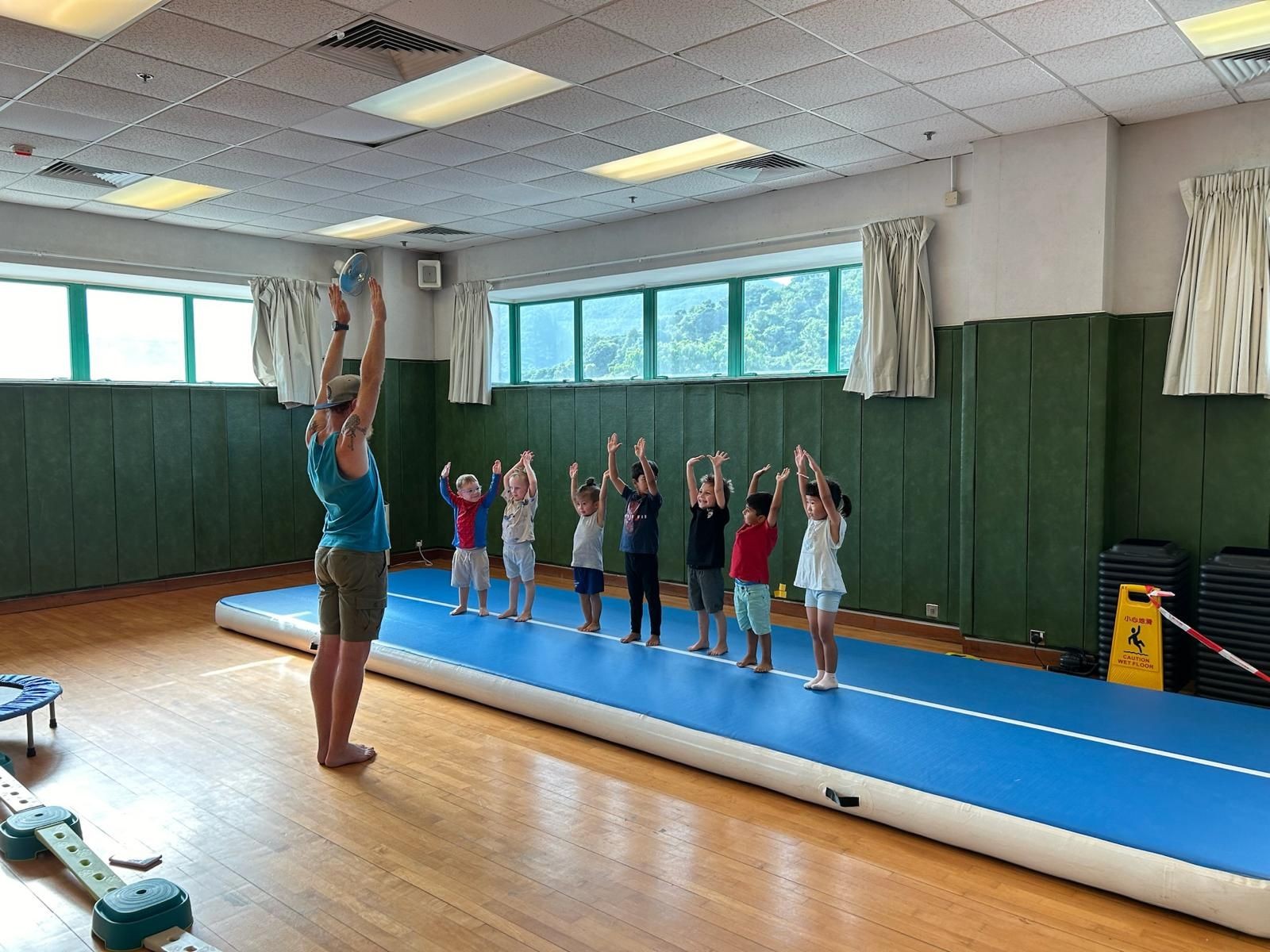 Children standing on a blue mat in a gym, raising their arms, led by an instructor.