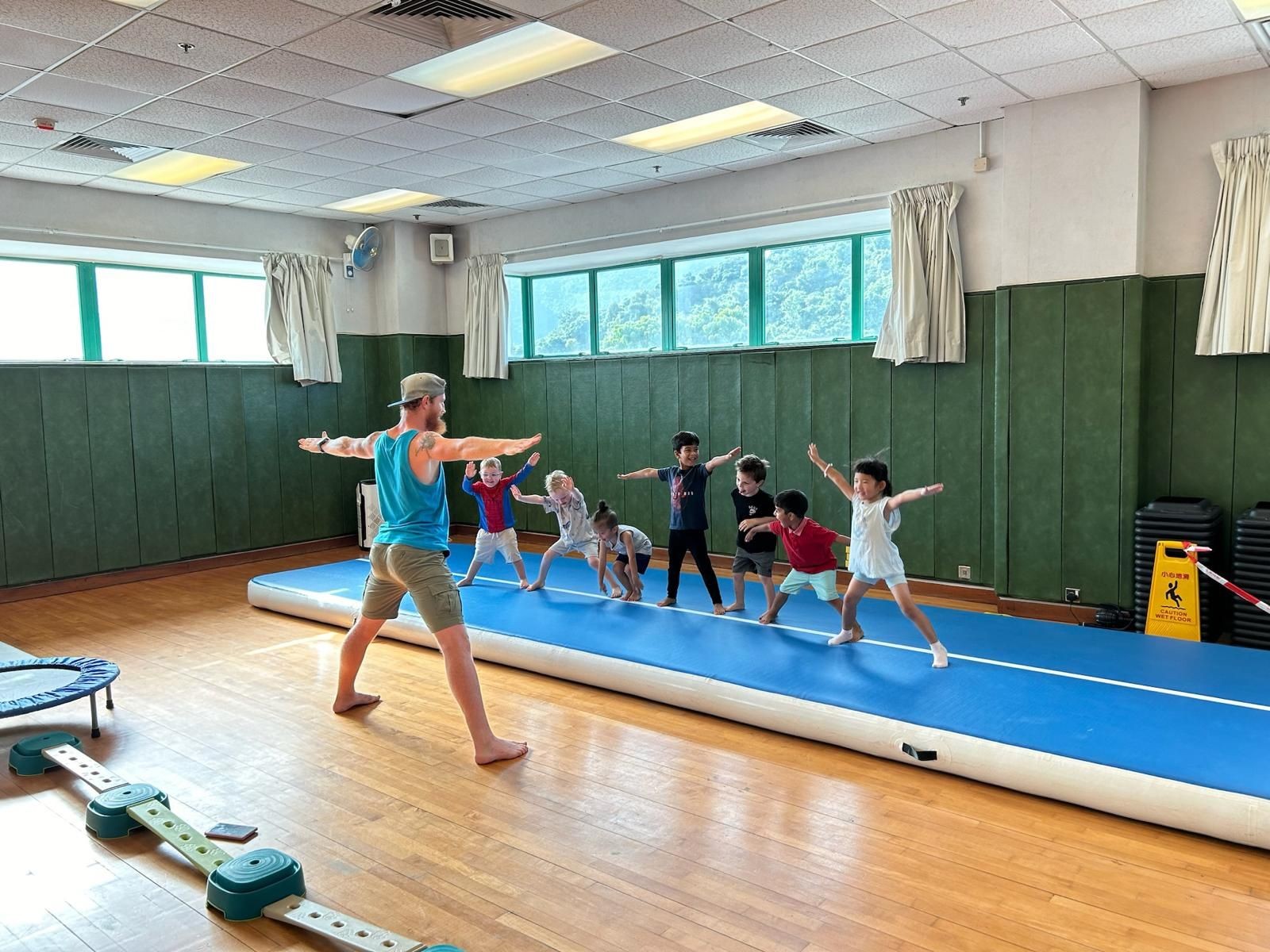 Instructor leading a group of children in a stretching exercise on a blue mat in a gymnasium.