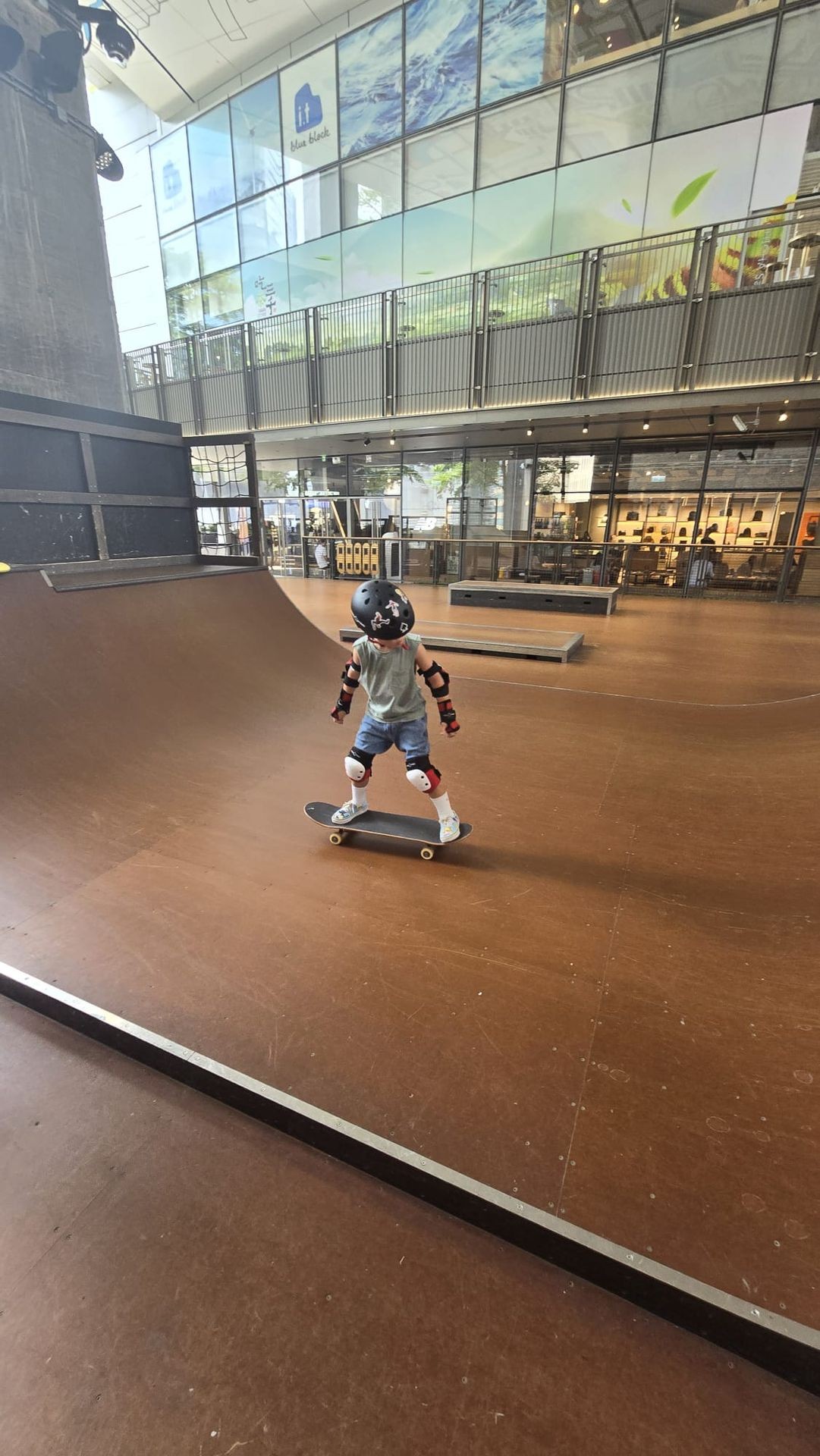 Child in protective gear skateboarding on an indoor ramp with glass windows in the background.