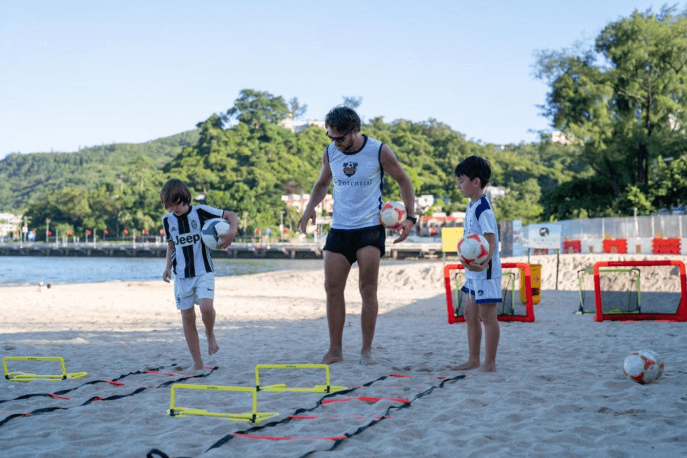 Coach and two kids practicing soccer drills on a sandy beach with small goals and hurdles.