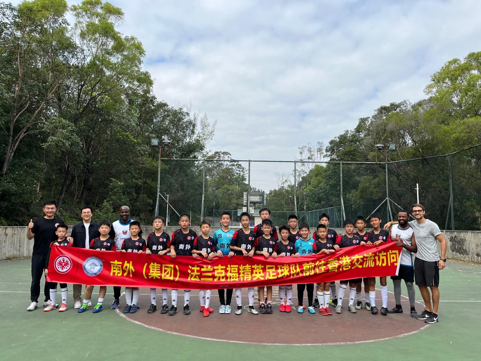 Group of young soccer players and coaches holding a red banner on an outdoor court surrounded by trees.