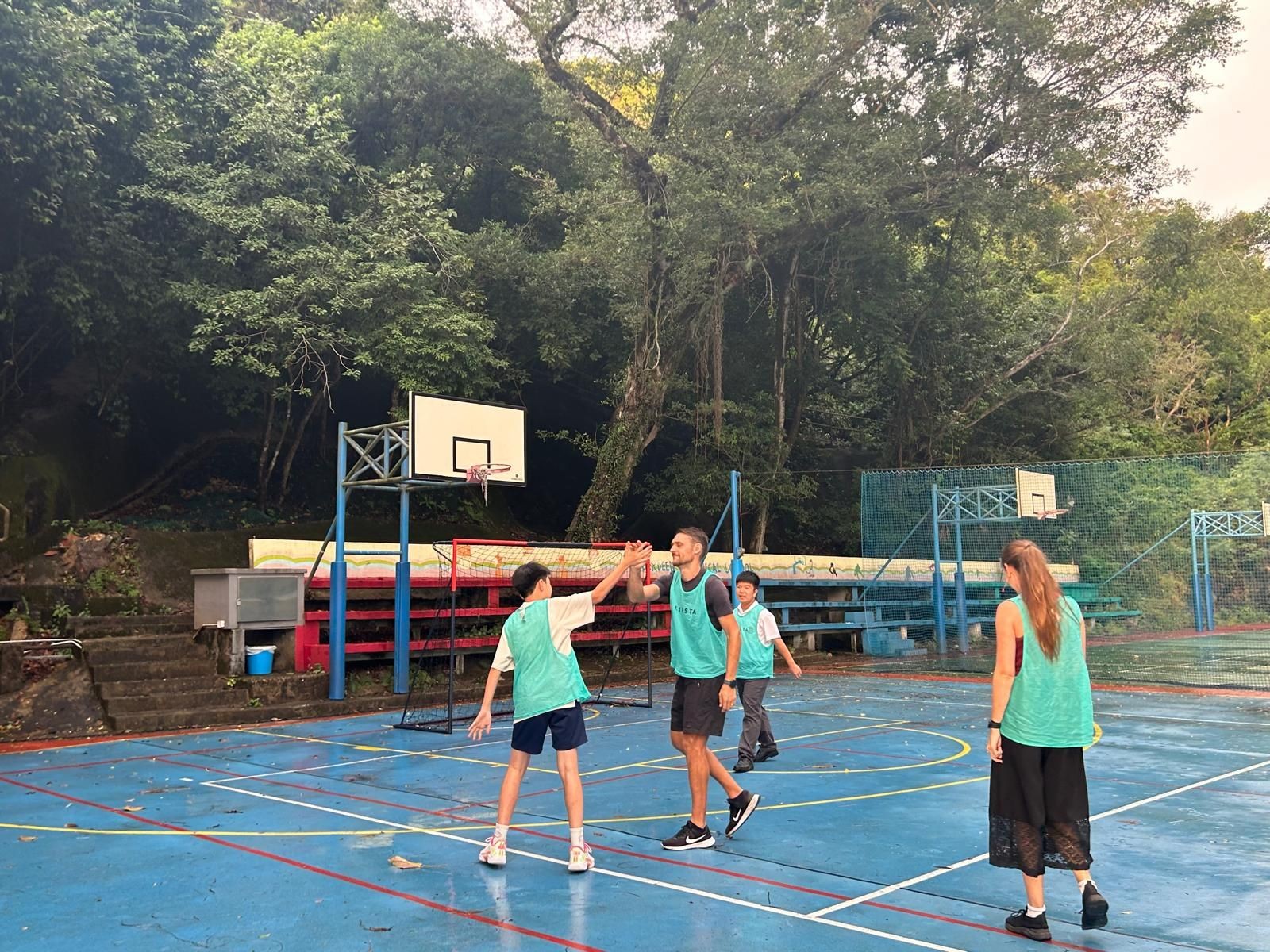 People playing basketball on an outdoor court surrounded by trees.